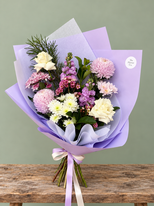 A stunning purple and white flower bouquet wrapped in tissue paper, tied with a pale purple ribbon. The bouquet contains white purple and pink flowers with green foliage. Flower delivery Adelaide. Flower delivery Golden Grove. The Flower Co. 