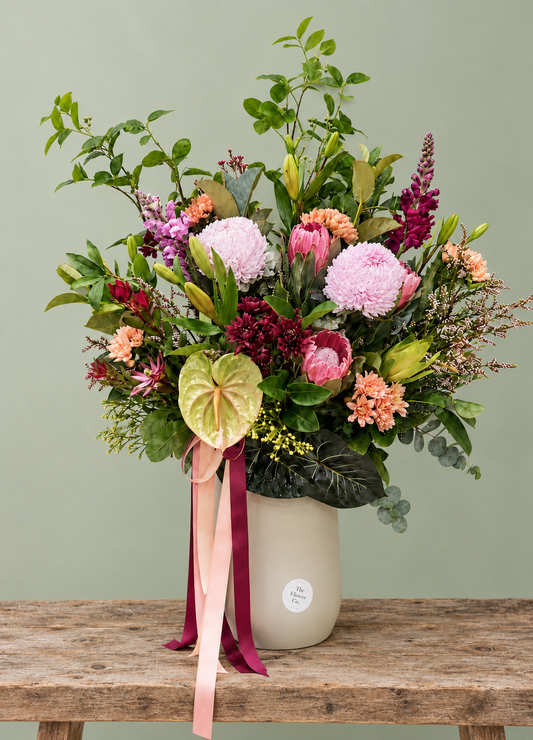 A vibrant mixed native flowers in a ceramic vase. The bouquet features a variety of flowers including pink chrysanthemums, burgundy protea, orange snapdragons, and green foliage, tied with pink and burgundy ribbons. Flower Delivery Adelaide. 